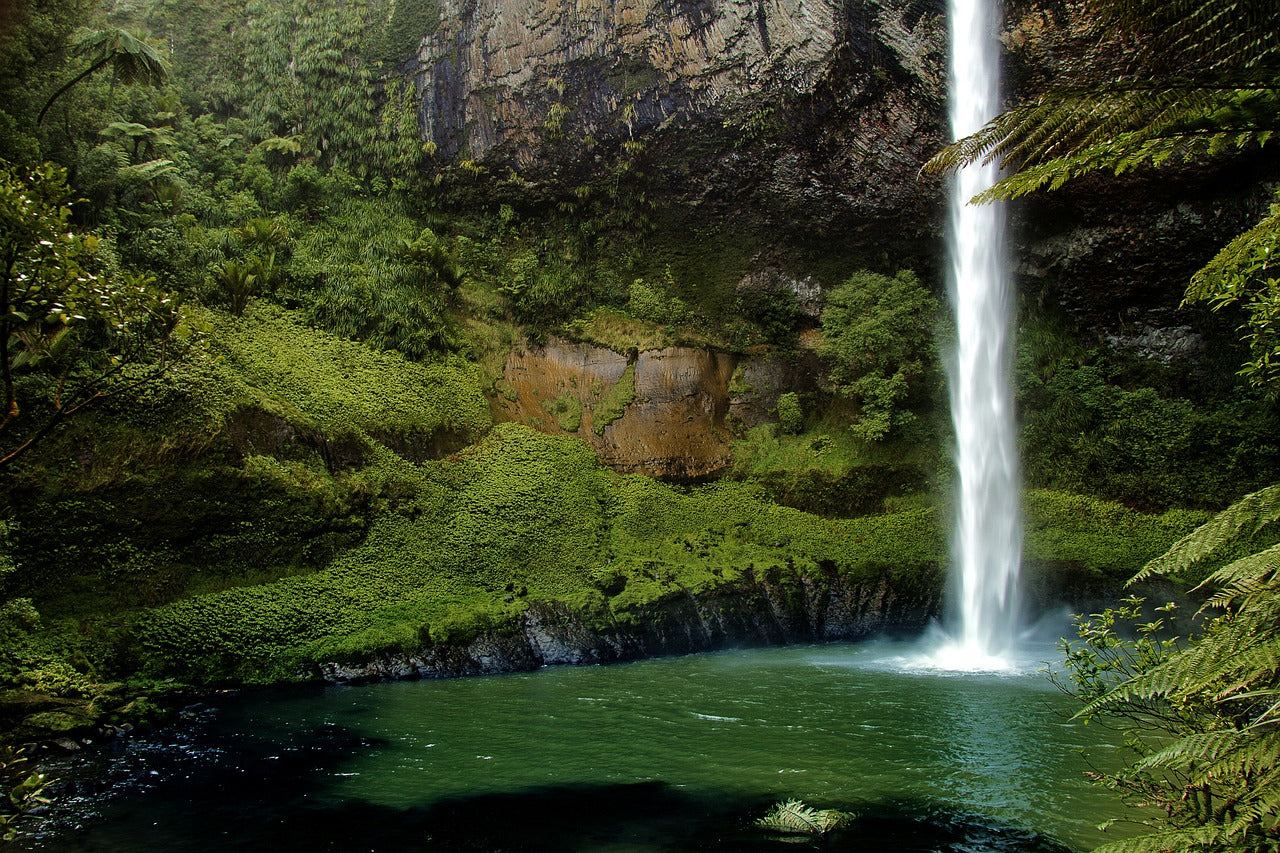 A thin water fall surrounded by green cliff faces and water pool.